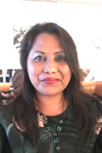 Lady with long dark hair, wearing pink lipstick, a green patterned top and smiling.