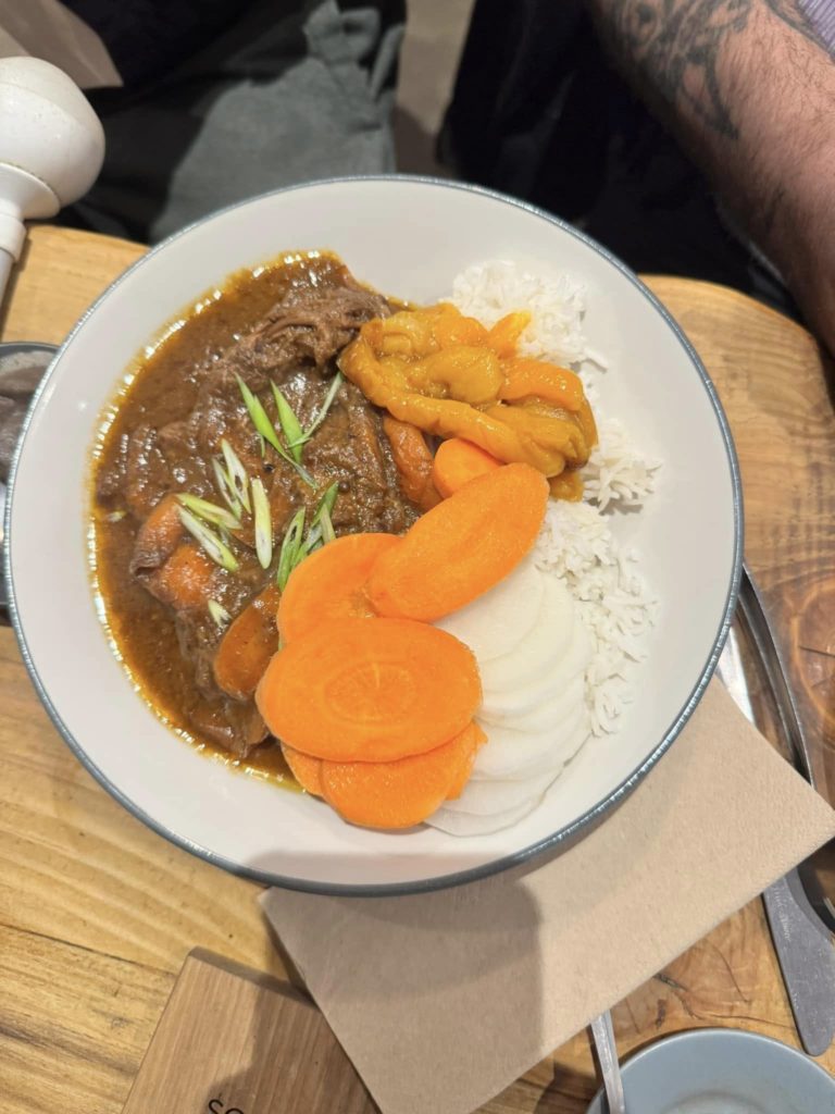 The image shows a meal which was available from the café. It is as bowl of braised beef which is in a brown sauce on a bed of white rice. The meal has been served with sliced carrot and marinated peppers.