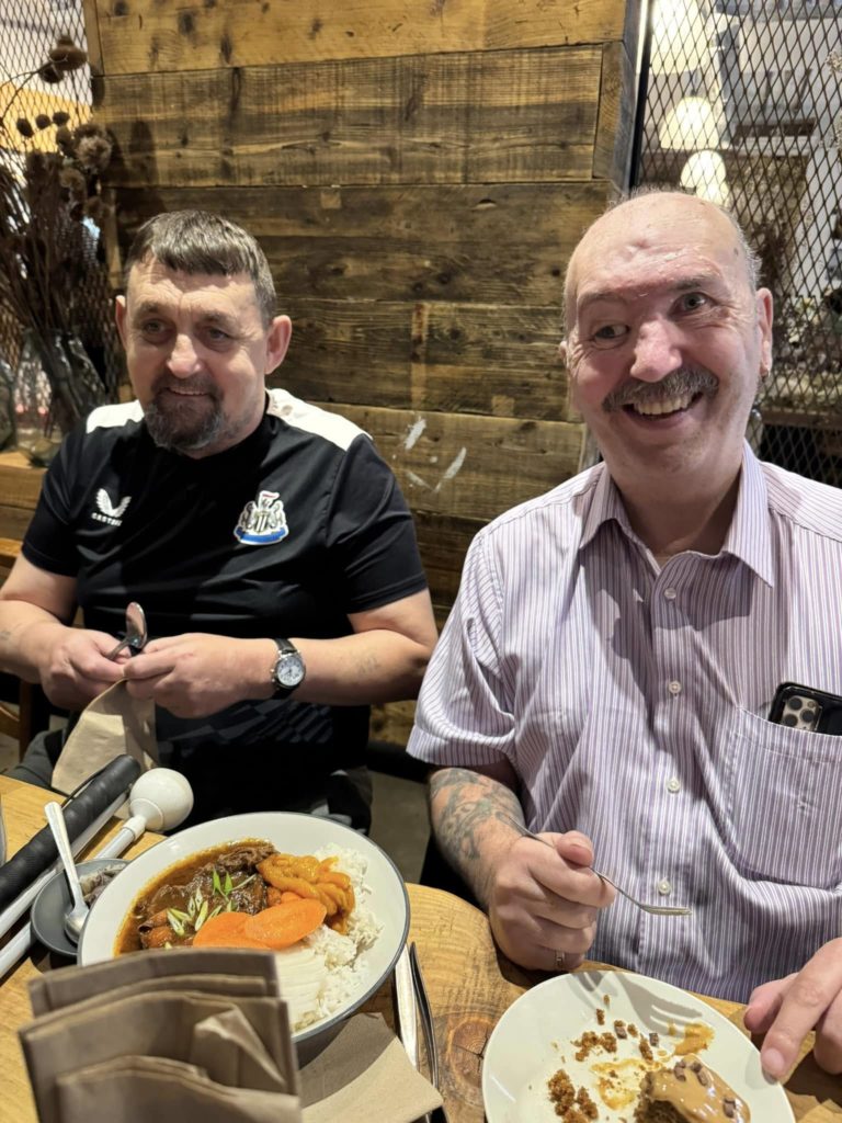 This image shows two men sat at a table, looking happy, with plates of food in front of them. One plate of food is a bowl of braised beef with carrots, rice and peppers. The other plate is carrot cake with toffee sauce and fudge pieces.