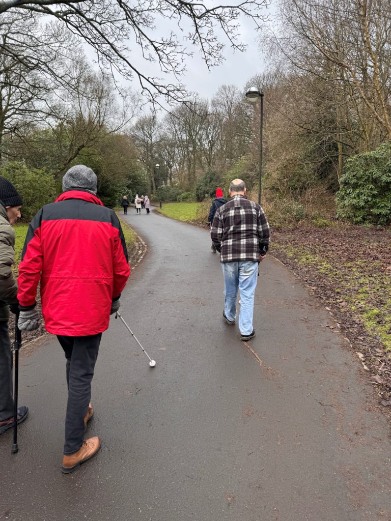 Image is of a man in a red coat walking with a long white cane. In front of him there is another man in blue jeans and a brown checked shirt. They are surrounded by green grass and trees with no leaves.