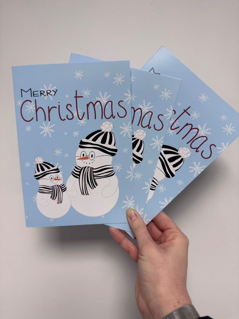 3 Christmas Cards being held up. The card is blue and there is a small snowman next to a larger snowman. The snowmen have carrot noses and are both wearing black and white scarves and black and white bobble hats. They are surrounded by white snowflakes. The text at the top of the card reads Merry Christmas.